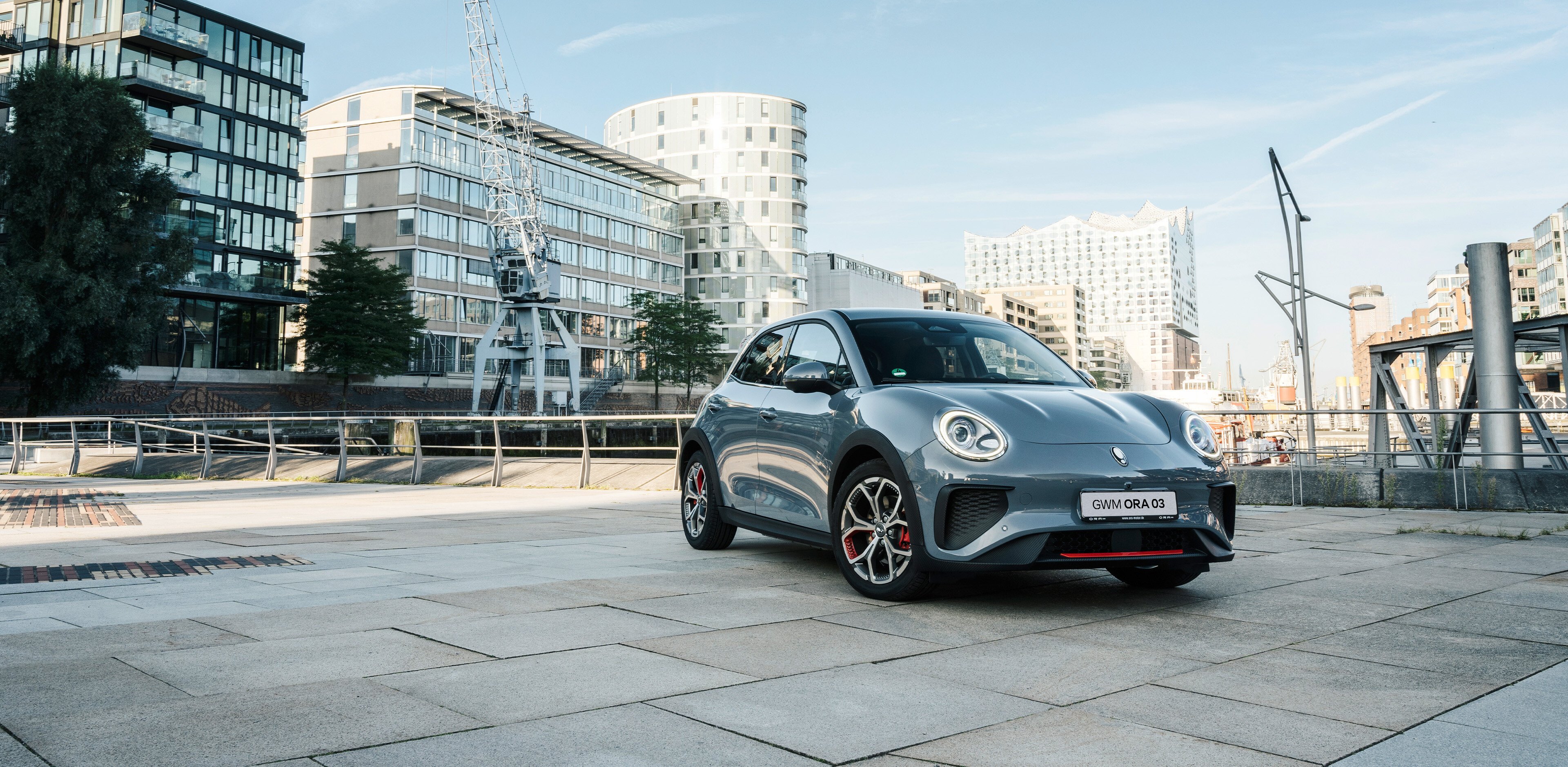 Der GWM ORA 03 GT in Meteorite Grey mit sportlichen Stoßfängern und Leichtmetallfelgen im GT Stil vor der Kulisse der Hamburger Elbphilharmonie