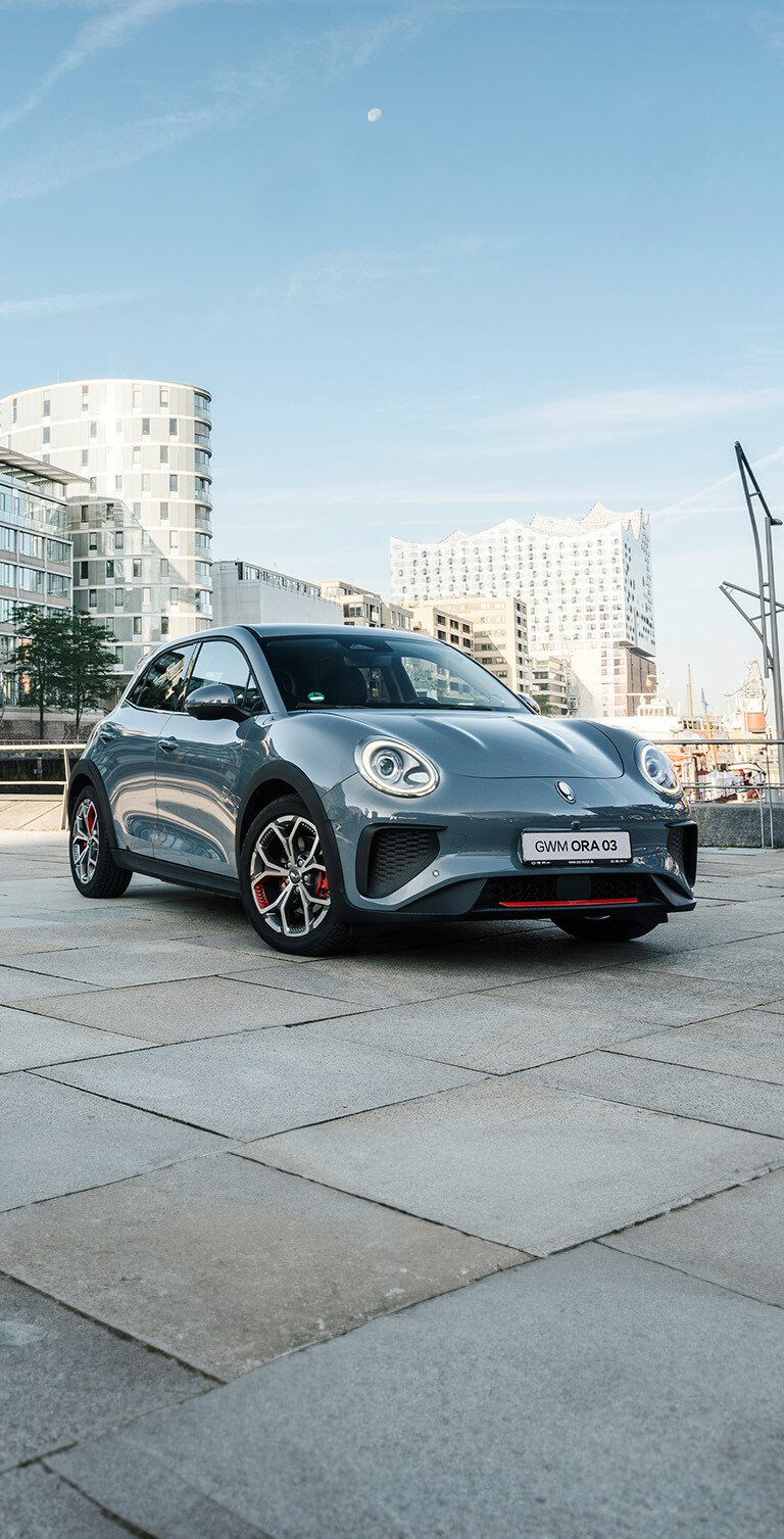 Der GWM ORA 03 GT in Meteorite Grey mit sportlichen Stoßfängern und Leichtmetallfelgen im GT Stil vor der Kulisse der Hamburger Elbphilharmonie