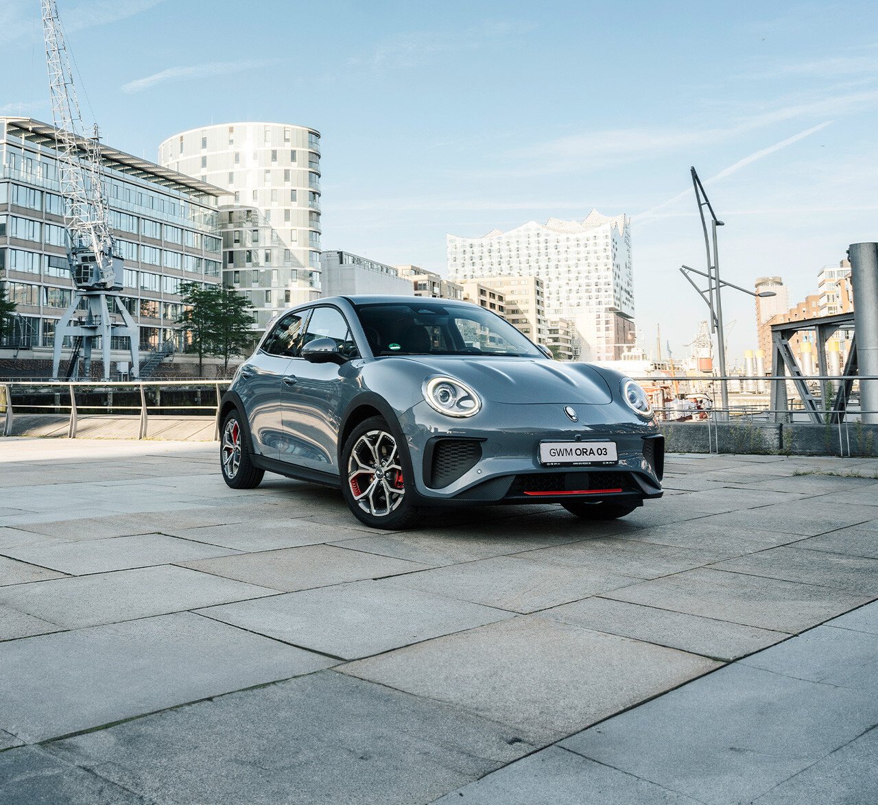 Der GWM ORA 03 GT in Meteorite Grey mit sportlichen Stoßfängern und Leichtmetallfelgen im GT Stil vor der Kulisse der Hamburger Elbphilharmonie