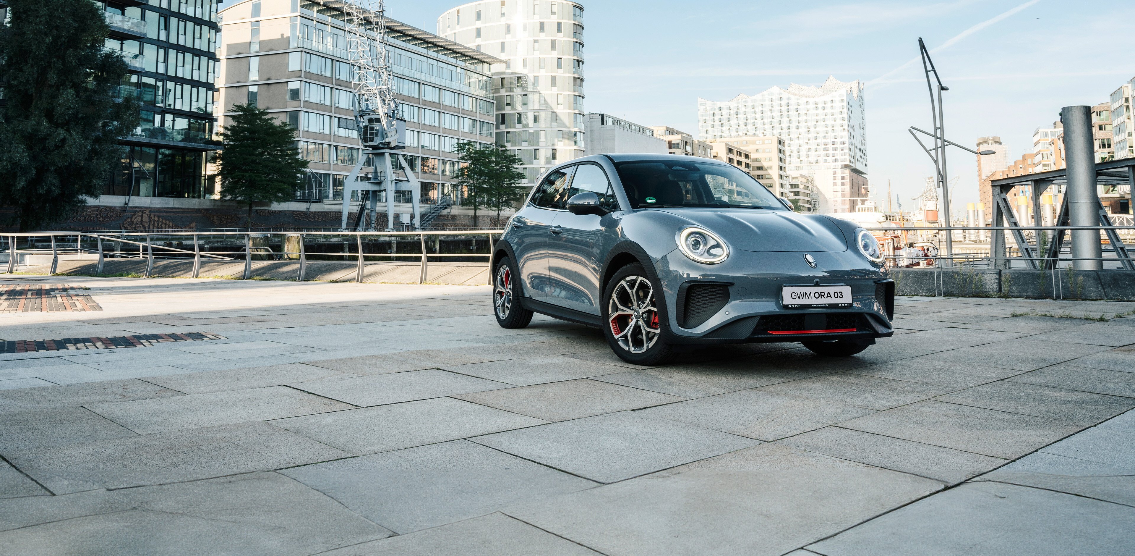 Der GWM ORA 03 GT in Meteorite Grey mit sportlichen Stoßfängern und Leichtmetallfelgen im GT Stil vor der Kulisse der Hamburger Elbphilharmonie