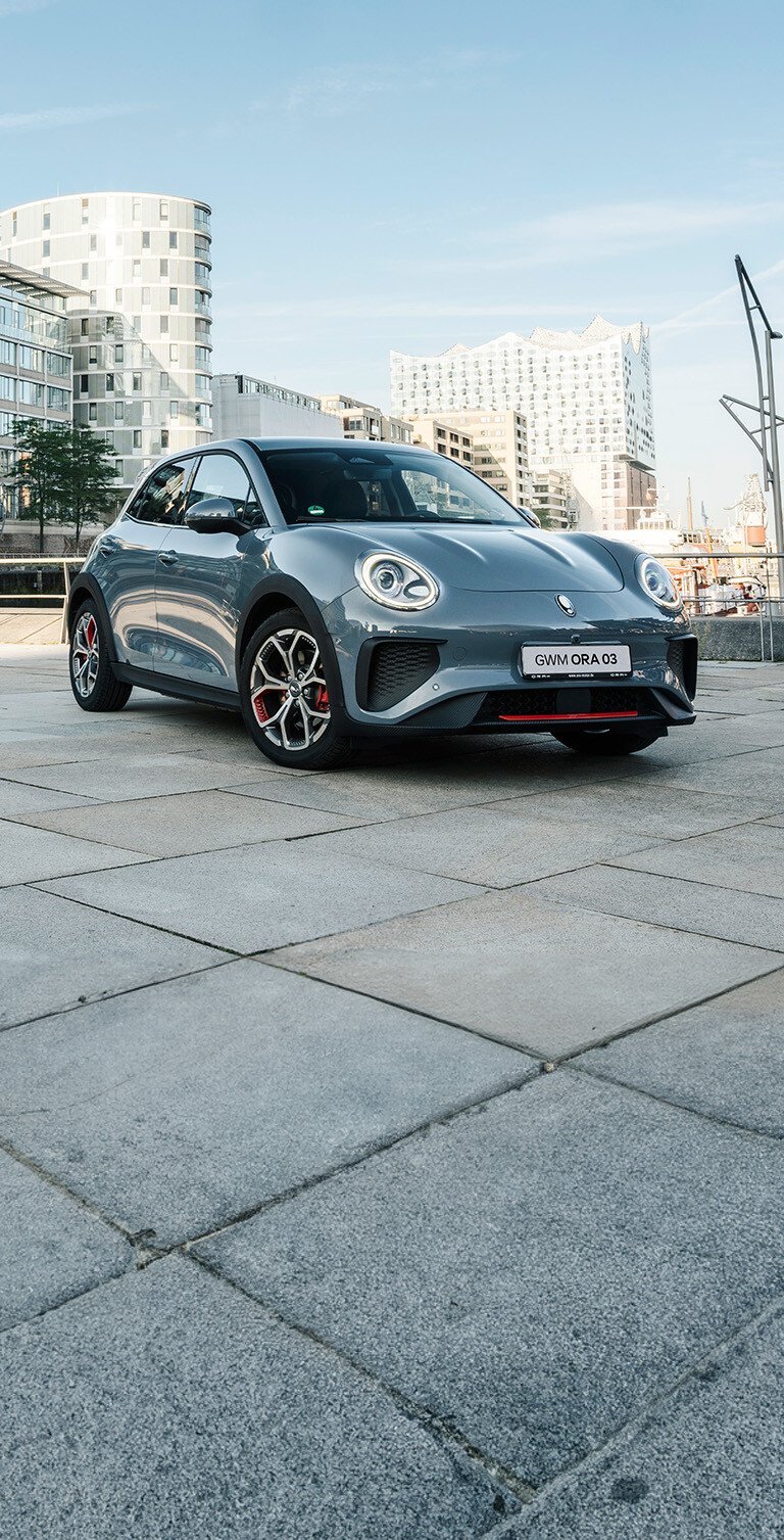 Der GWM ORA 03 GT in Meteorite Grey mit sportlichen Stoßfängern und Leichtmetallfelgen im GT Stil vor der Kulisse der Hamburger Elbphilharmonie